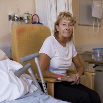 A blonde haired woman sits in a chair next to a hospital bed and a pair of crutches.